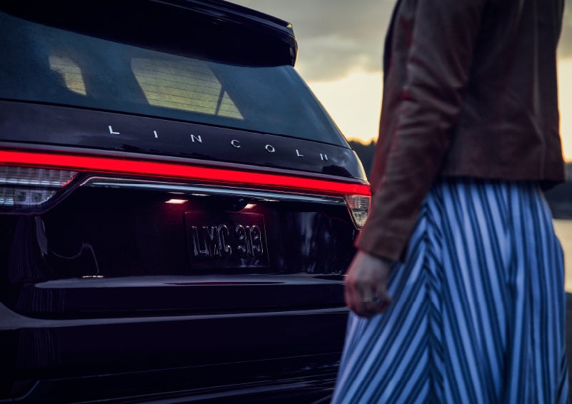 A person is shown near the rear of a 2024 Lincoln Aviator® SUV as the Lincoln Embrace illuminates the rear lights | Lincoln of Cutler Bay in Miami FL