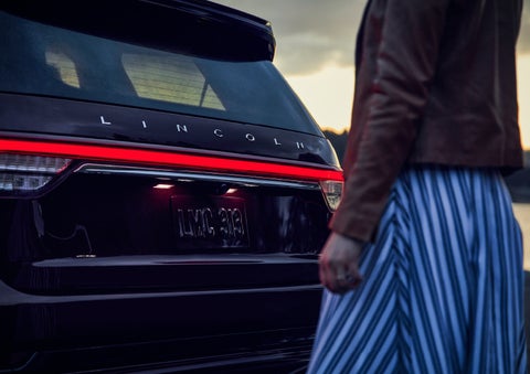 A person is shown near the rear of a 2024 Lincoln Aviator® SUV as the Lincoln Embrace illuminates the rear lights | Lincoln of Cutler Bay in Miami FL